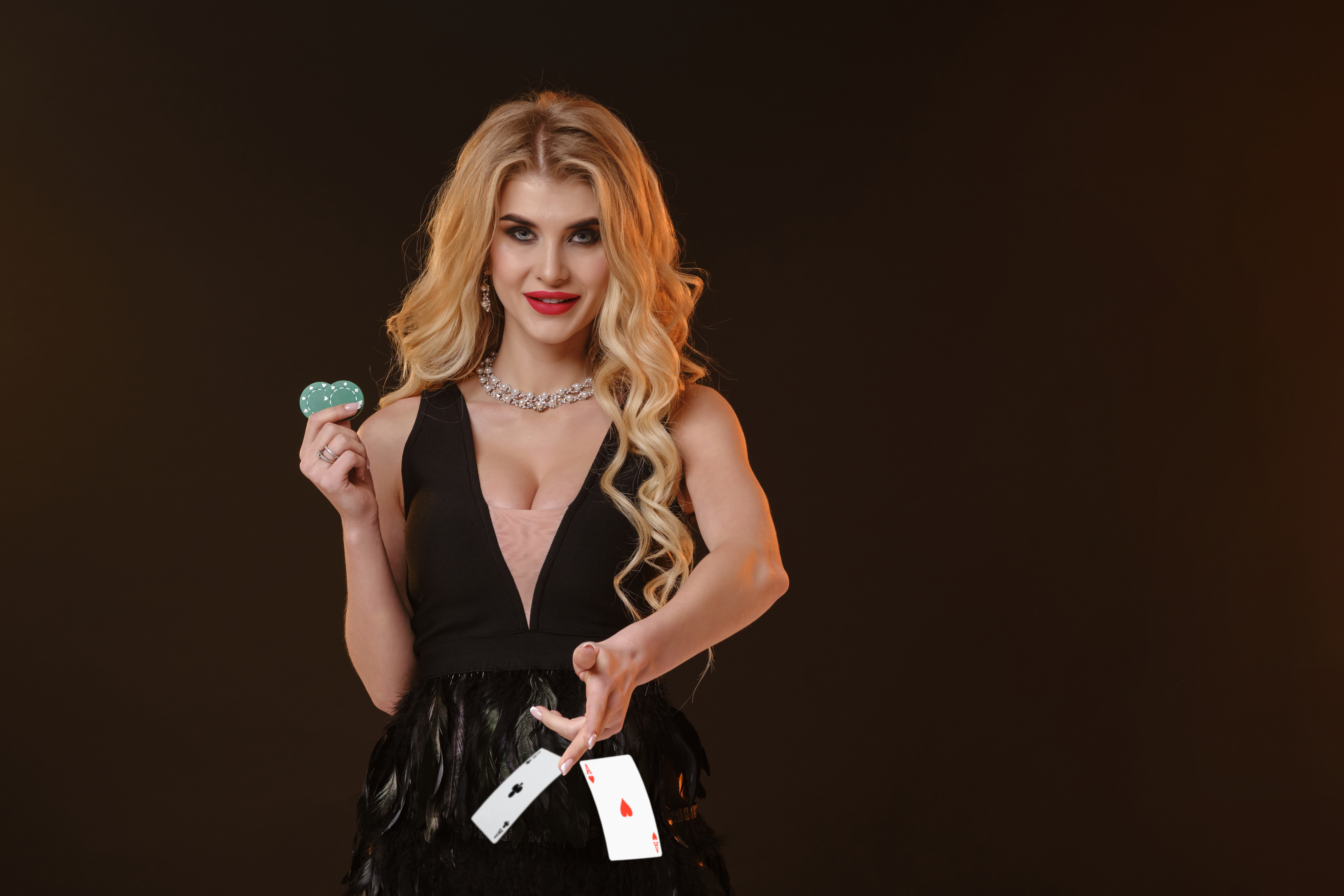 Blonde girl in black dress and necklace. Smiling, showing two green chips, throwing something, posing on brown background. Poker, casino. Close-up